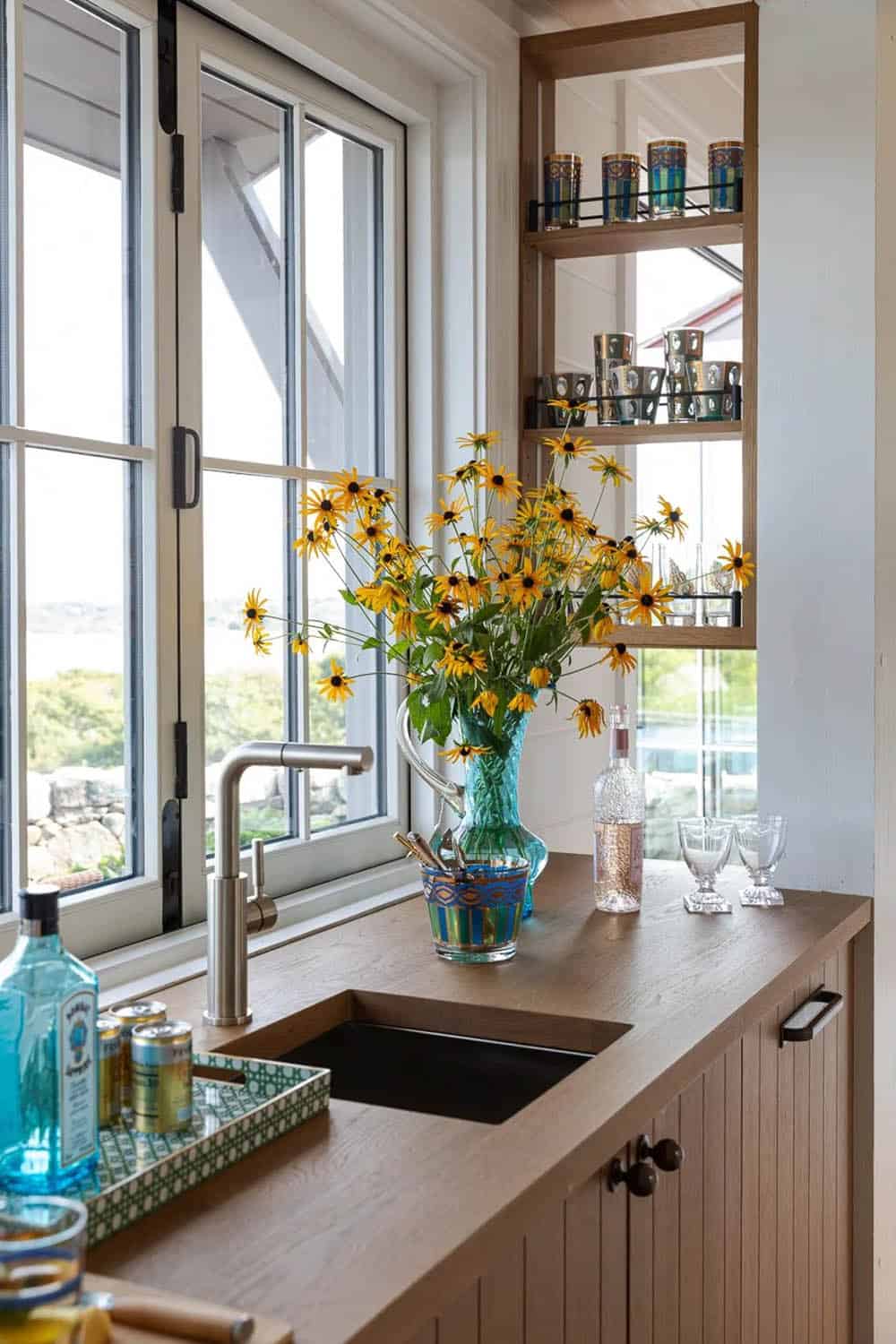 Wet bar counter with black sink, wildflowers, and glassware shelving by window