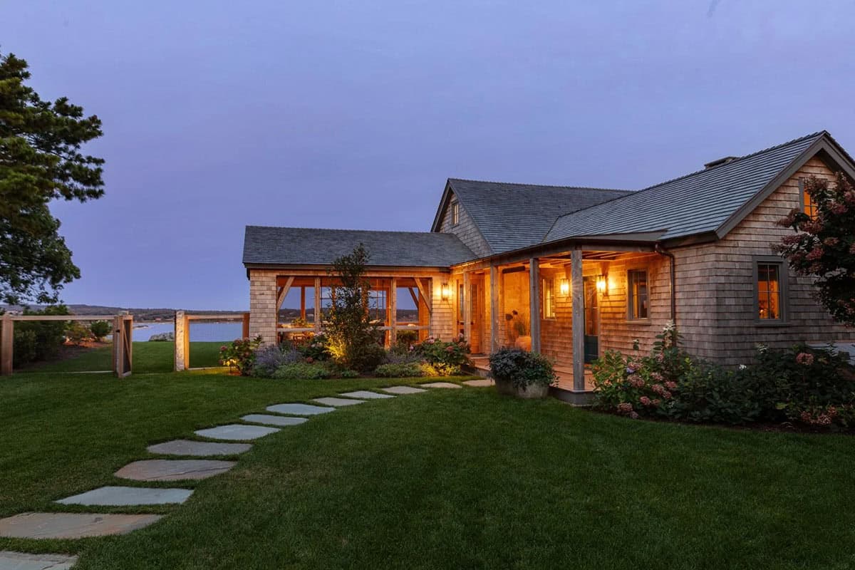 Twilight view of shingle-style home with glowing porch lights and stone pathway to lawn