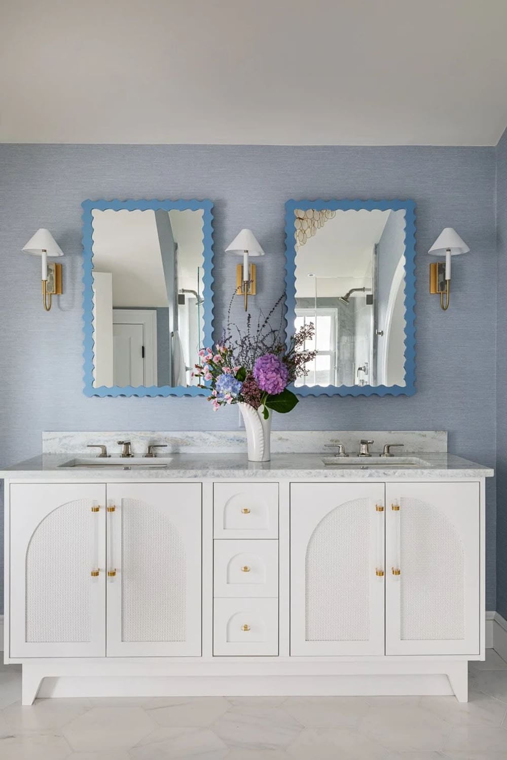 Double vanity bathroom with scalloped blue mirrors and brass sconces on blue grasscloth wall
