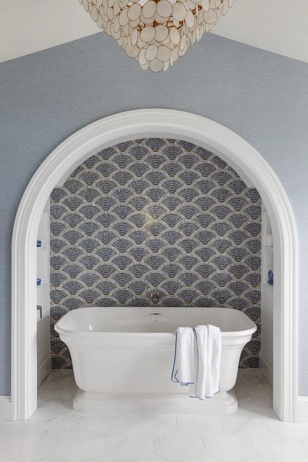 Freestanding tub under capiz shell chandelier framed by arched fan-tile alcove