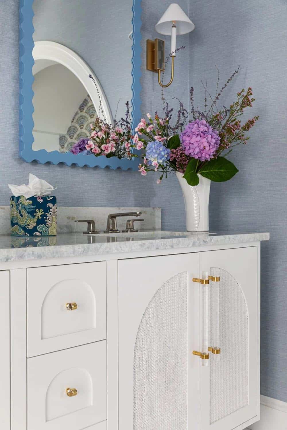 Bathroom vanity close-up with scalloped blue mirror, brass sconce, and hydrangea bouquet
