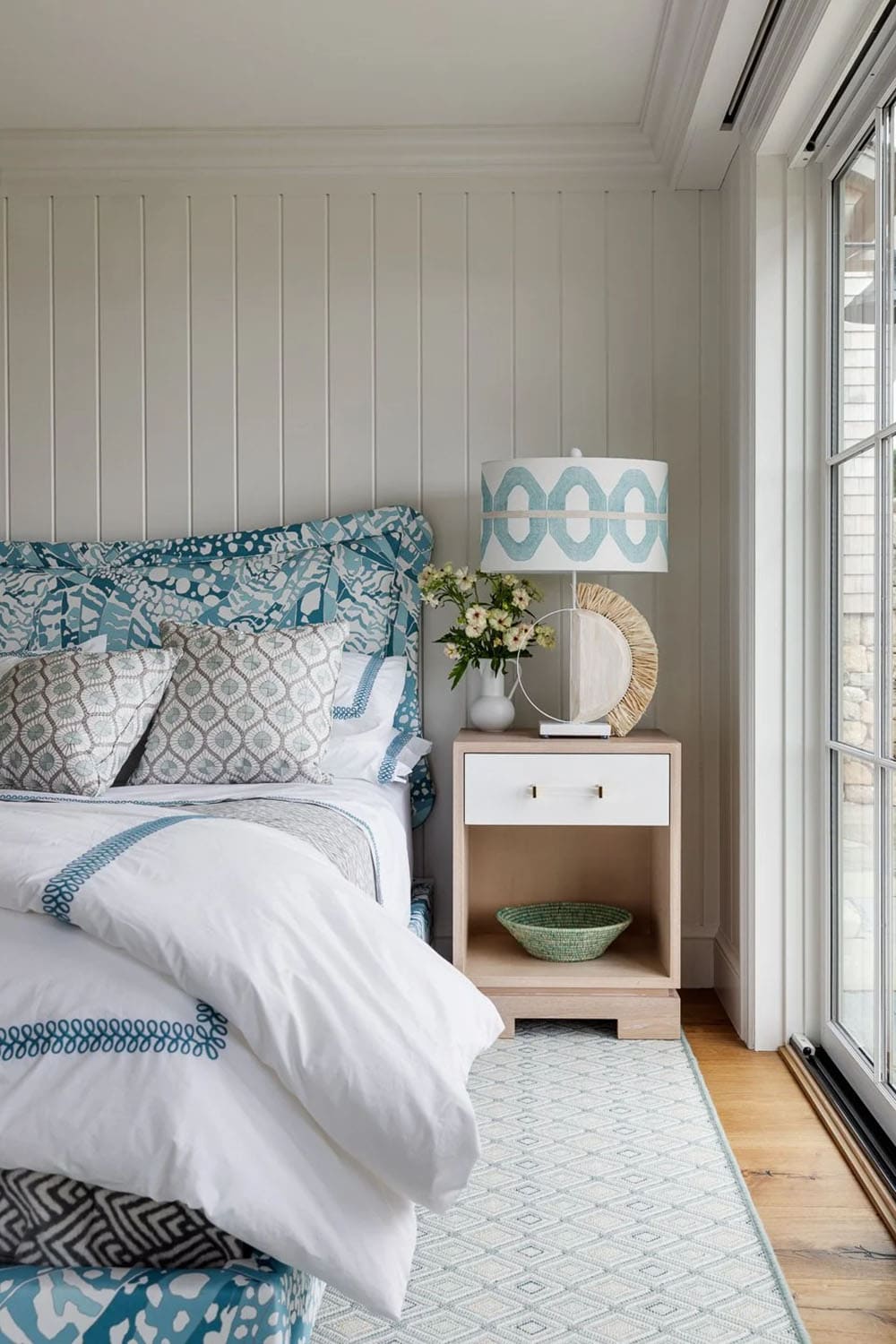 Bedroom nightstand with teal patterned lamp, white bedding, and diamond-pattern rug