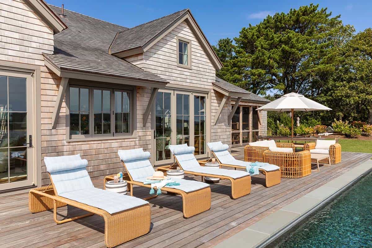Pool deck lined with wicker chaise lounges beside shingled home with folding glass doors
