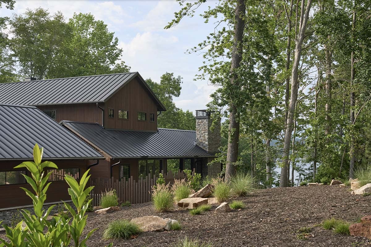 Rear exterior of a contemporary lake house with dark standing-seam metal roof, wood siding, and stone chimney, surrounded by trees