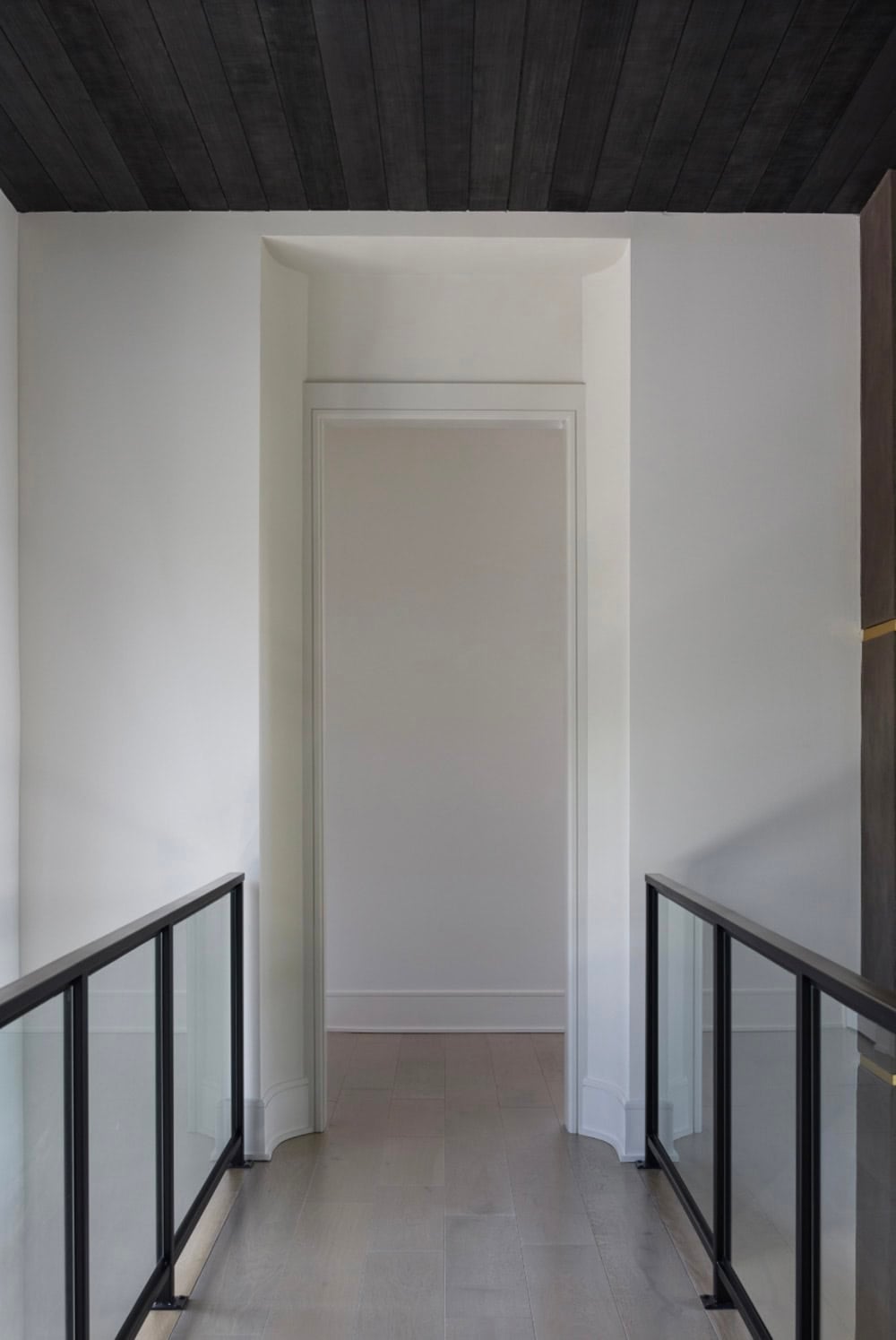 Upstairs hallway with dark wood ceiling, glass-and-black railing, and doorway to a bedroom