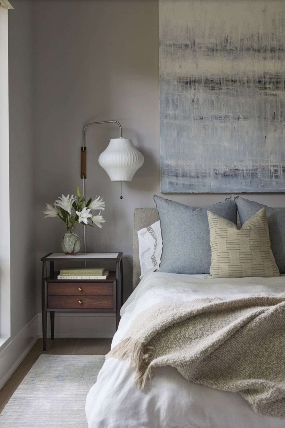 Bedside vignette with wood nightstand, ceramic lamp, fresh flowers, and abstract art above the bed
