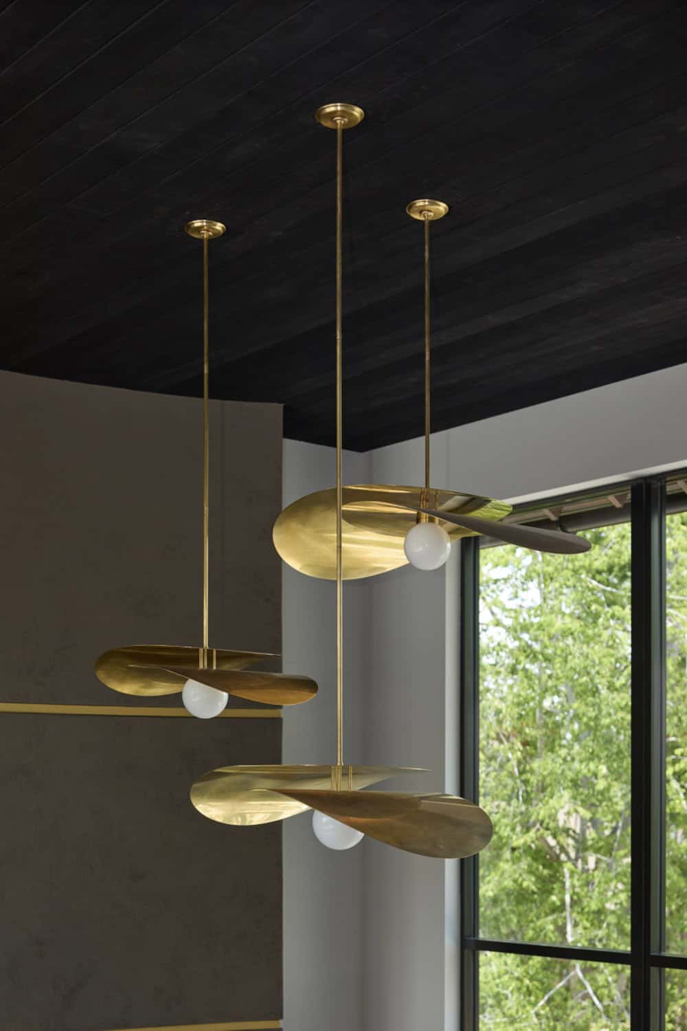 Sculptural brass pendant lights with disc shades hanging from a black wood ceiling near a window