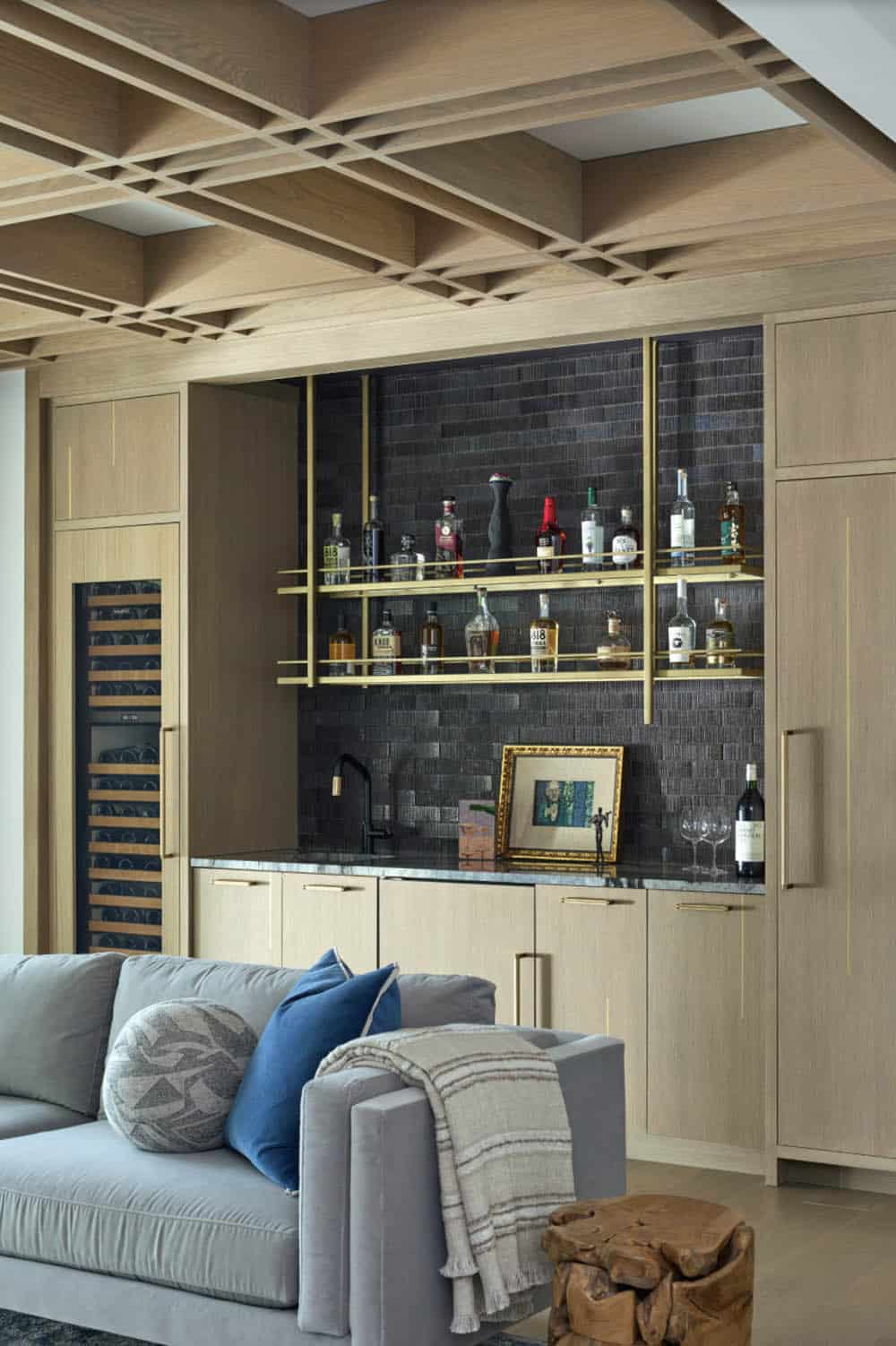Home bar with oak cabinetry, brass shelving, dark tile backsplash, and stocked liquor bottles