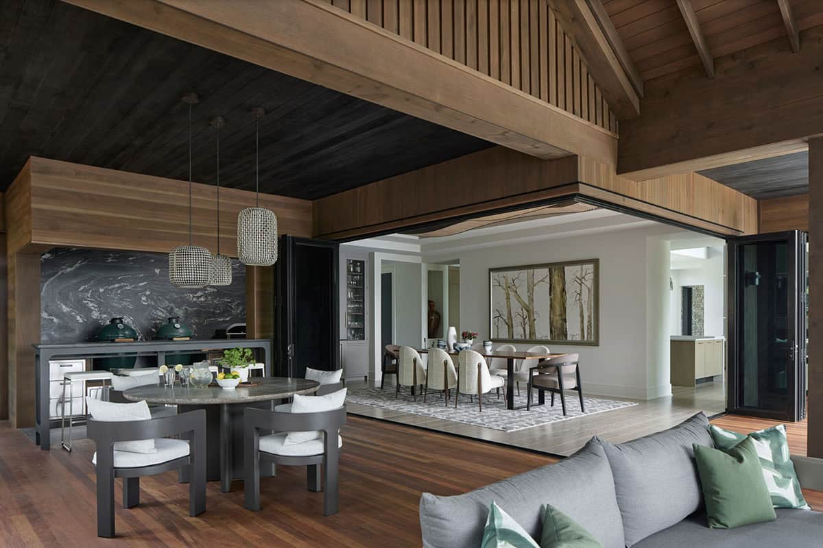 Open-air outdoor kitchen and dining area with dark stone counters, grills, and pendant lighting leading to an indoor dining room