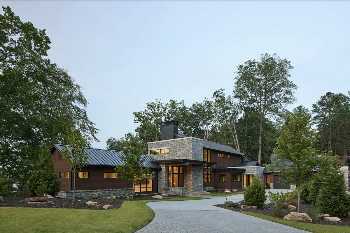 Dusk view of the house exterior with illuminated windows, stone chimney, and paver driveway