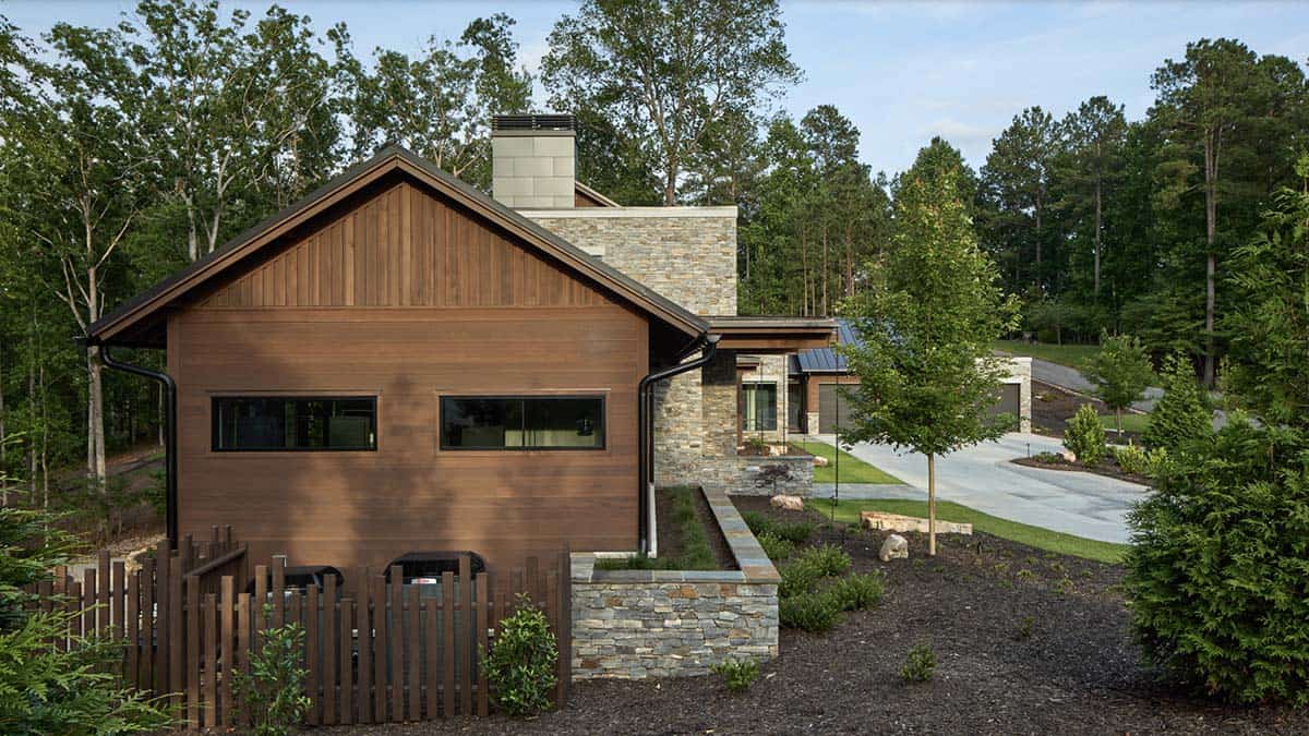 Side exterior view of wood-clad gable end, stone chimney, and mechanical enclosure behind a wood fence