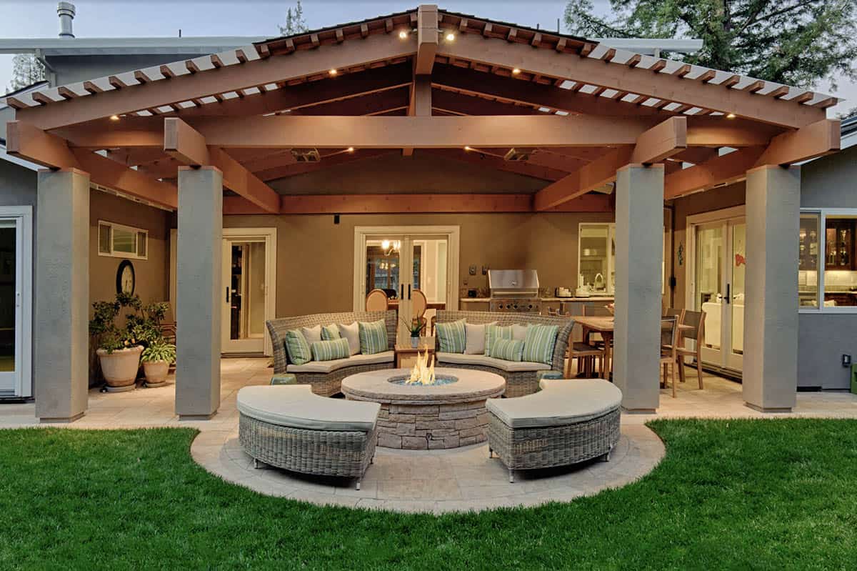 Covered patio with a large wood pergola-style roof, curved wicker seating around a round stone fire pit, and an outdoor kitchen with a grill and dining table visible in the background