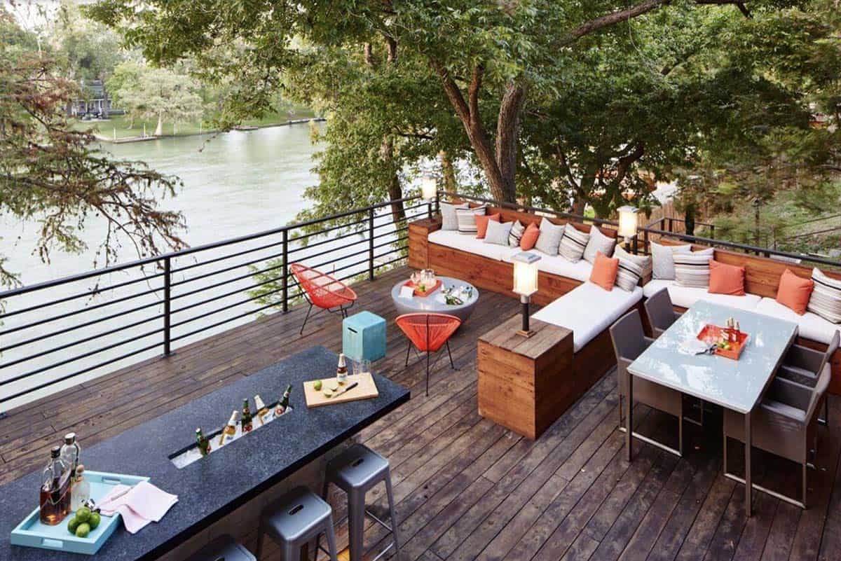 Elevated wood deck overlooking a river, with a stone bar counter, L-shaped sectional seating, a dining table, and orange accent chairs tucked among mature trees
