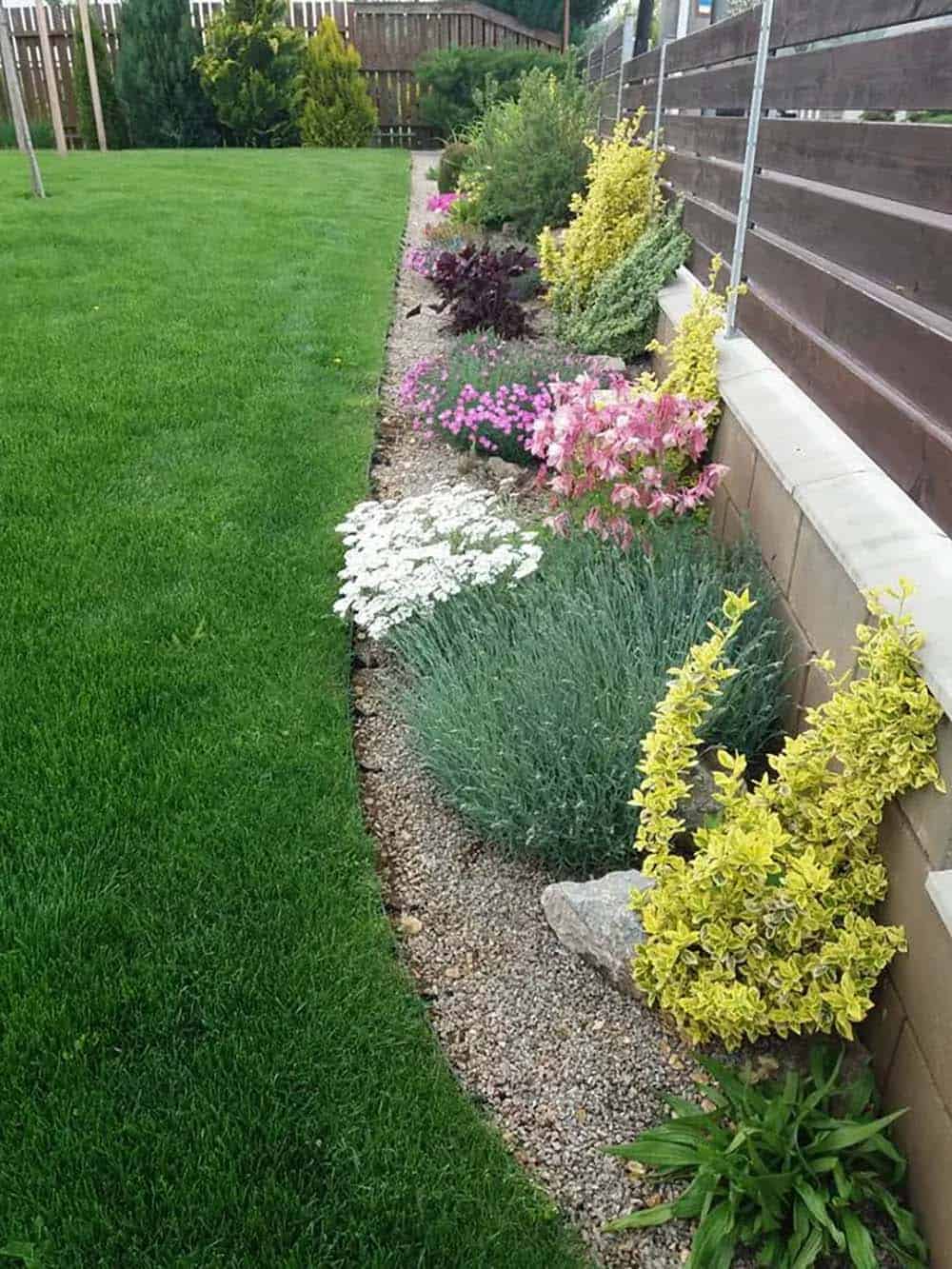 Gravel-lined border with colorful shrubs and flowers along a dark horizontal fence