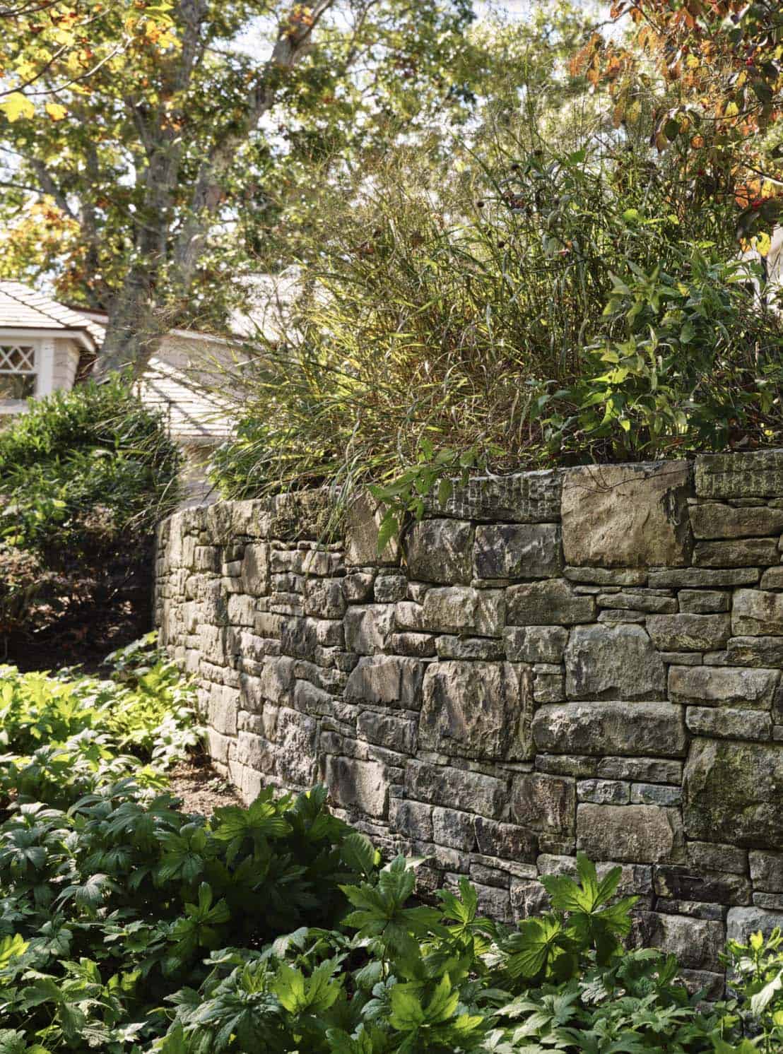 Dry-stacked stone retaining wall with garden plantings and glimpse of a shingled roof
