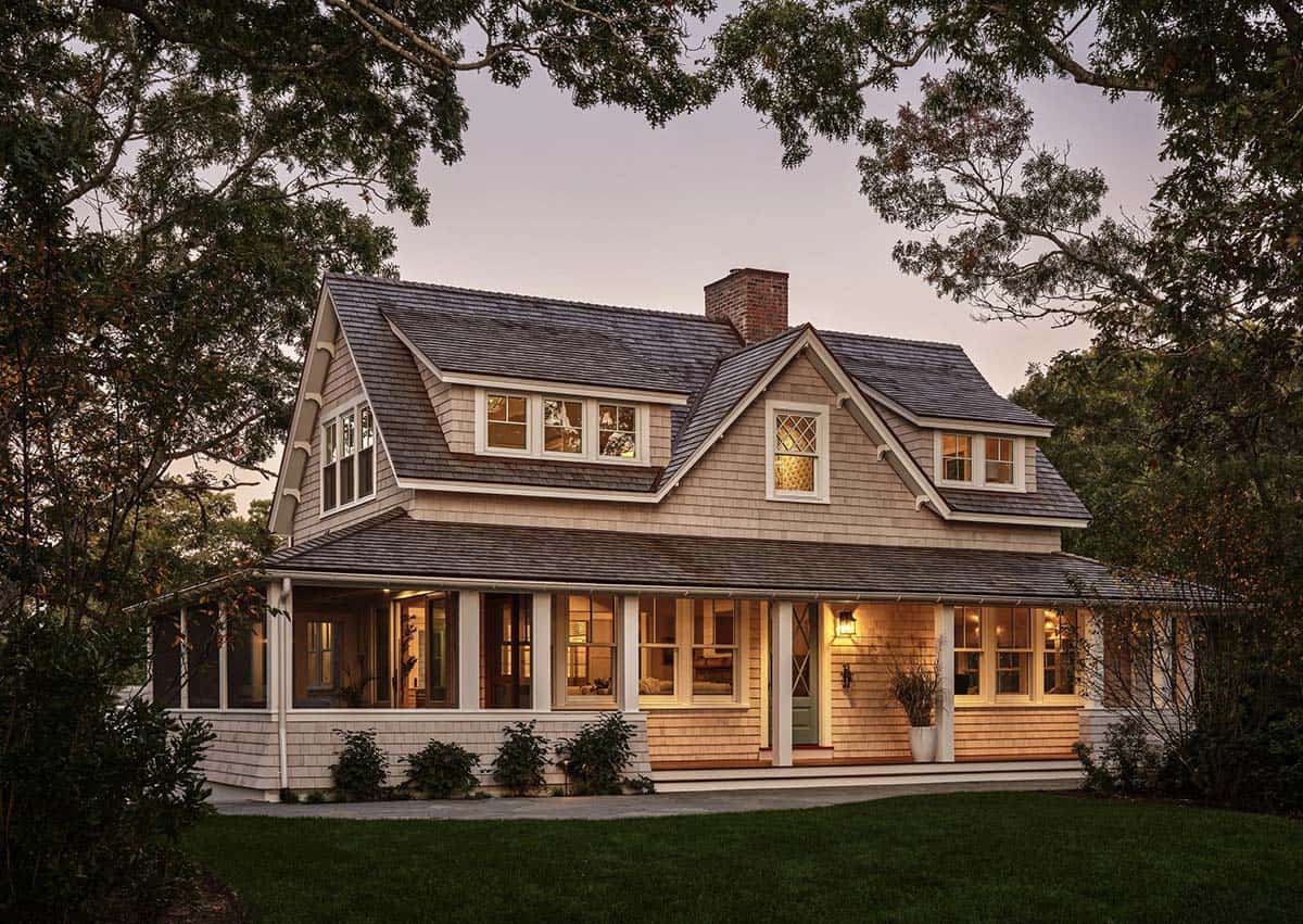 Twilight exterior of gambrel-roofed cottage with wraparound porch and warm interior lighting