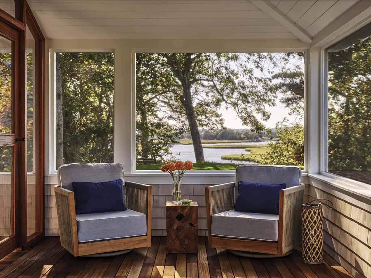 Screened porch with woven teak armchairs, navy pillows, and live-edge side table overlooking a marsh and pond