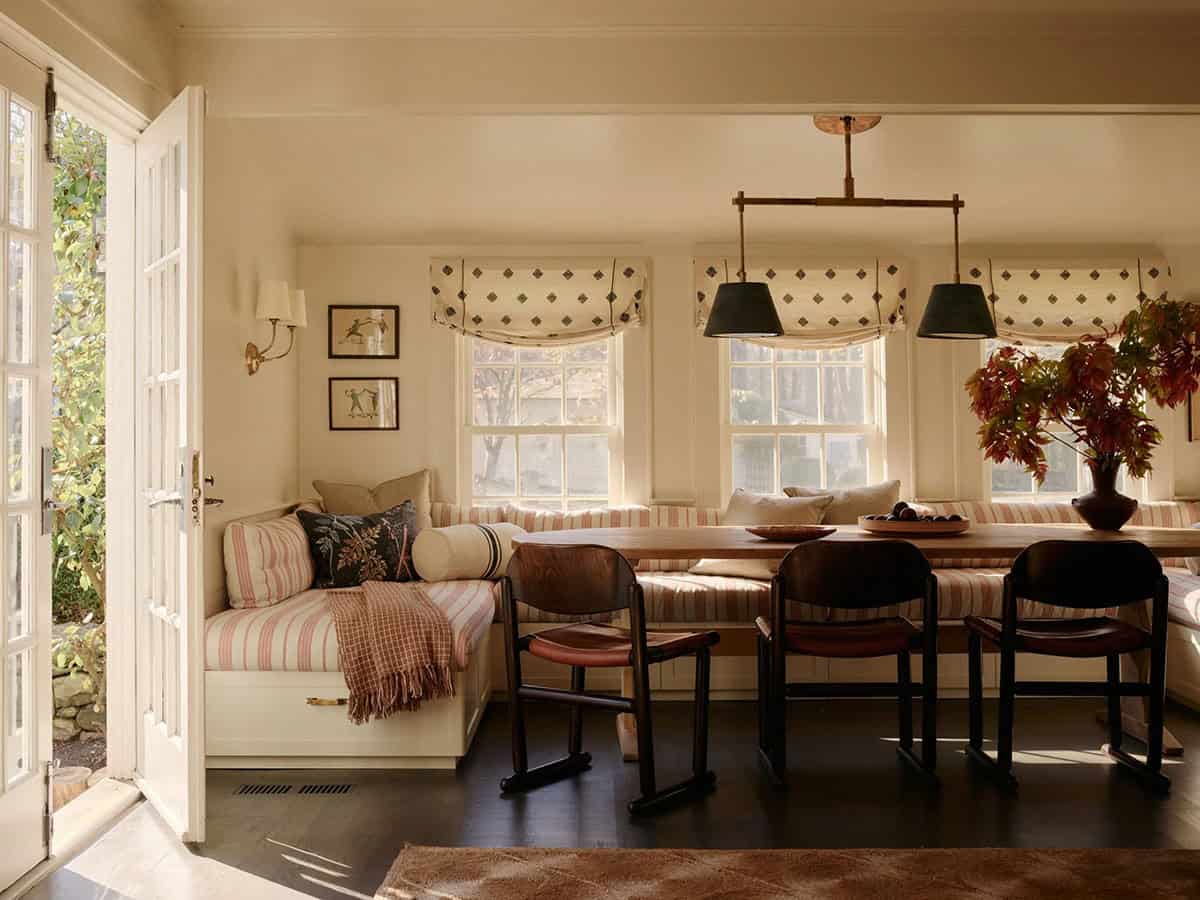 Breakfast nook with striped banquette, wood table, black chairs, and pendant lights by French doors