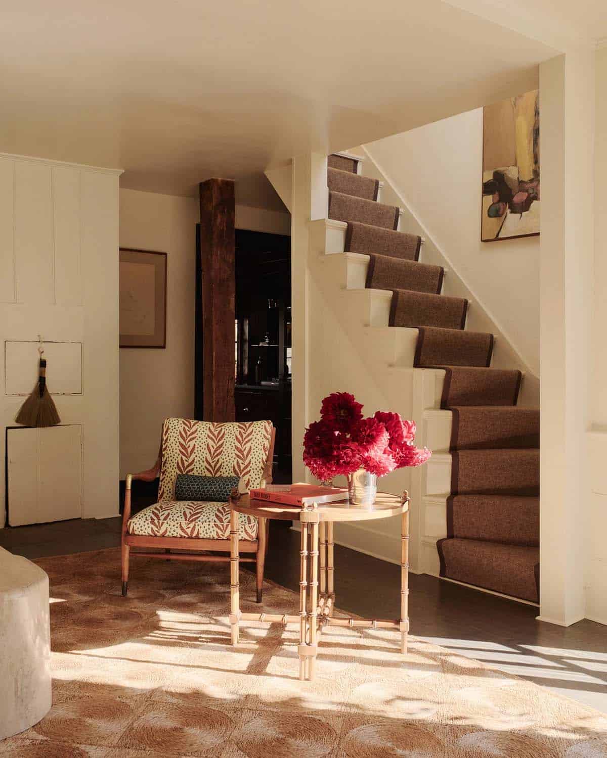 Entryway with patterned armchair, bamboo side table, red dahlias, and carpeted stairs