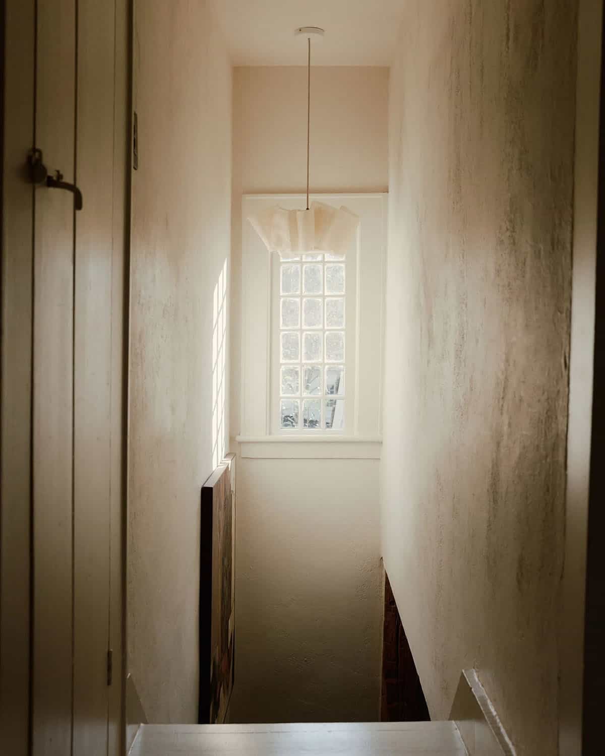 Narrow staircase landing with tall window, paper pendant light, mottled plaster walls