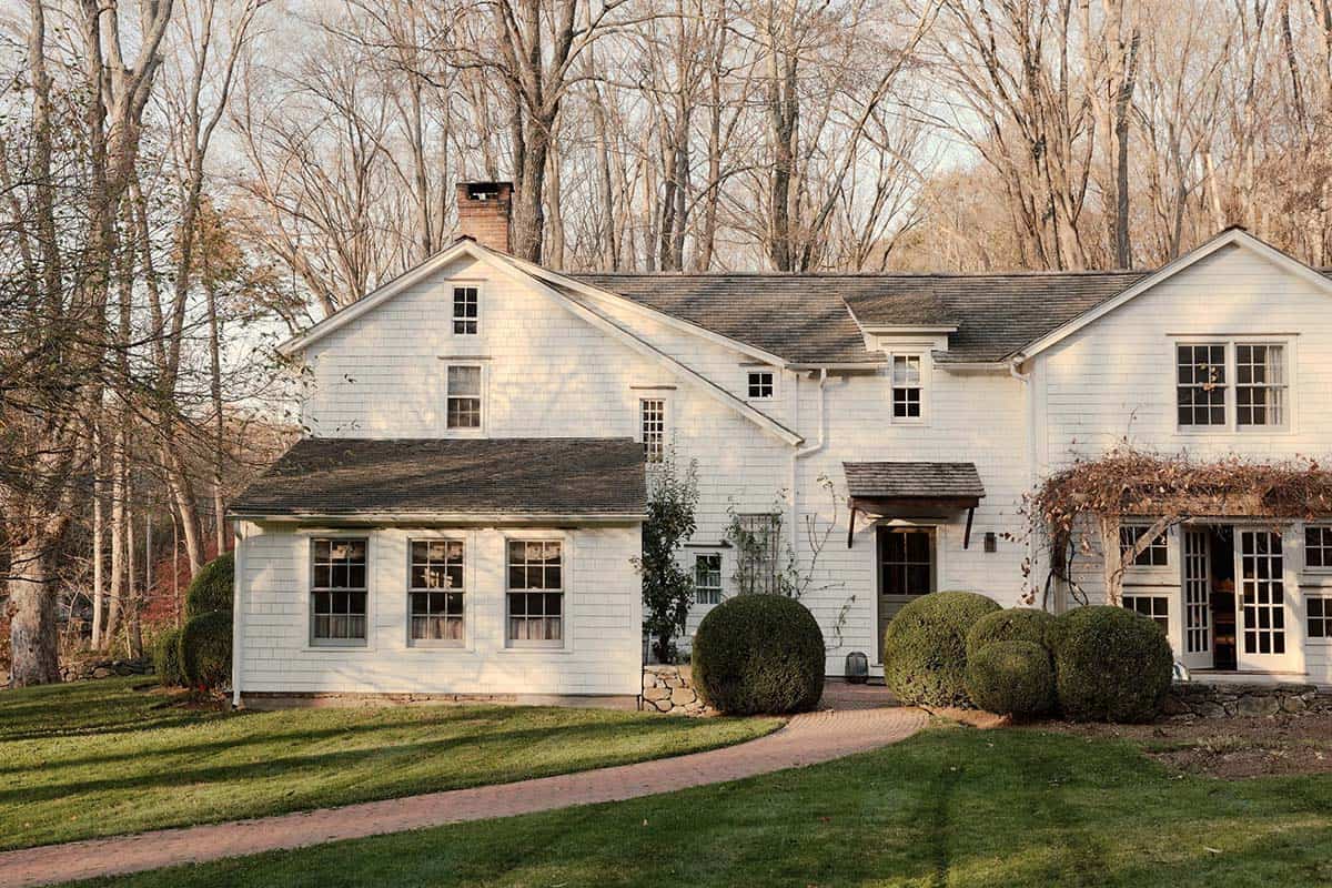 Exterior of white shingled farmhouse with brick chimney and boxwood shrubs