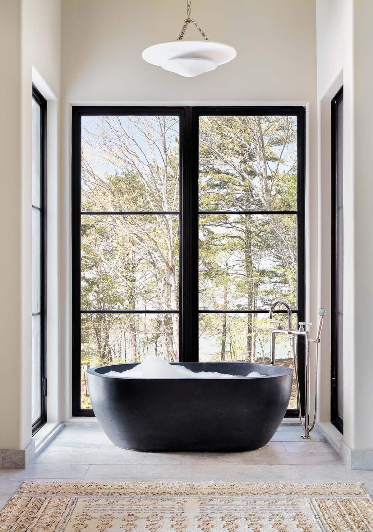Freestanding black soaking tub beneath tall forest facing windows