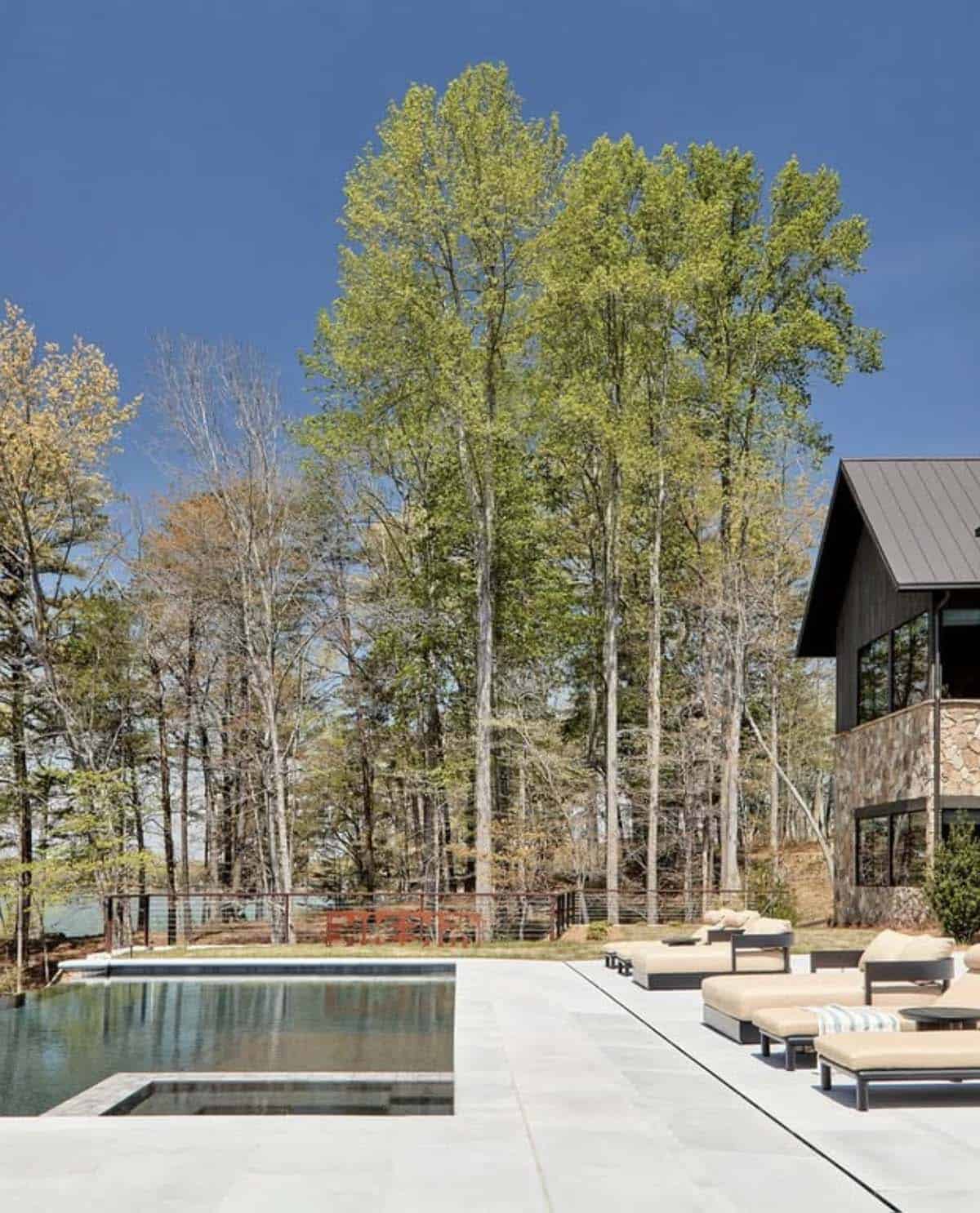 Poolside patio with lounge chairs beneath tall trees near lake house