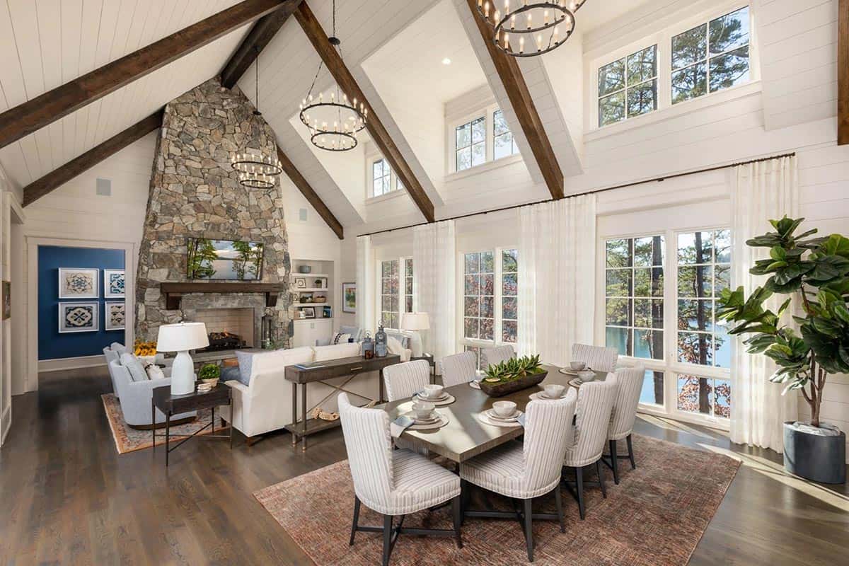 Vaulted great room with stone fireplace, wood beams, and dining table by lake-view windows