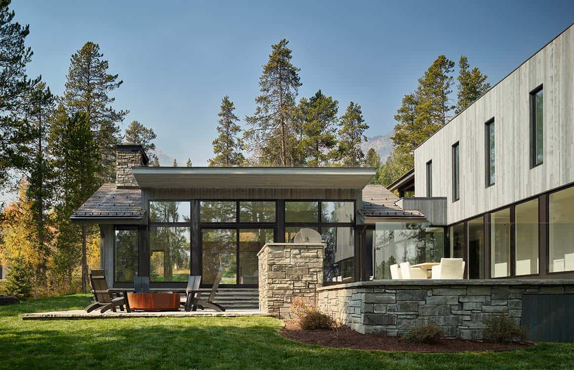 Rear exterior of contemporary mountain home with stone chimney, glass walls, and fire pit patio