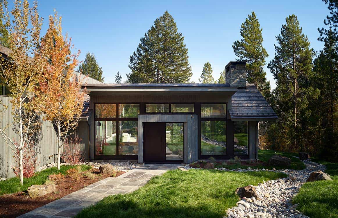 Front entrance with dark pivot door, floor-to-ceiling windows, and stone pathway through native landscaping