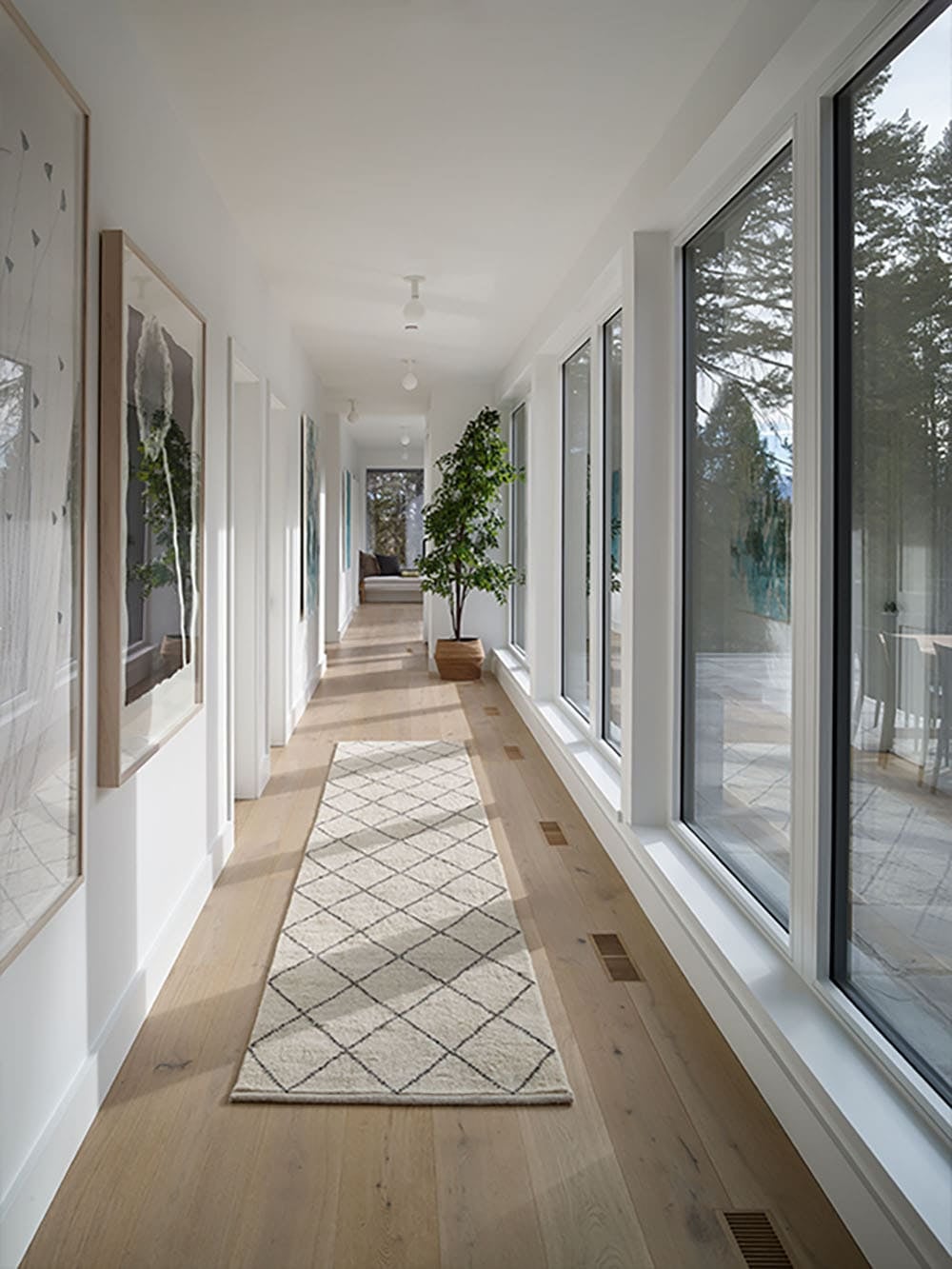 Bright hallway lined with framed art and floor-to-ceiling windows, leading past a potted tree to a sitting nook