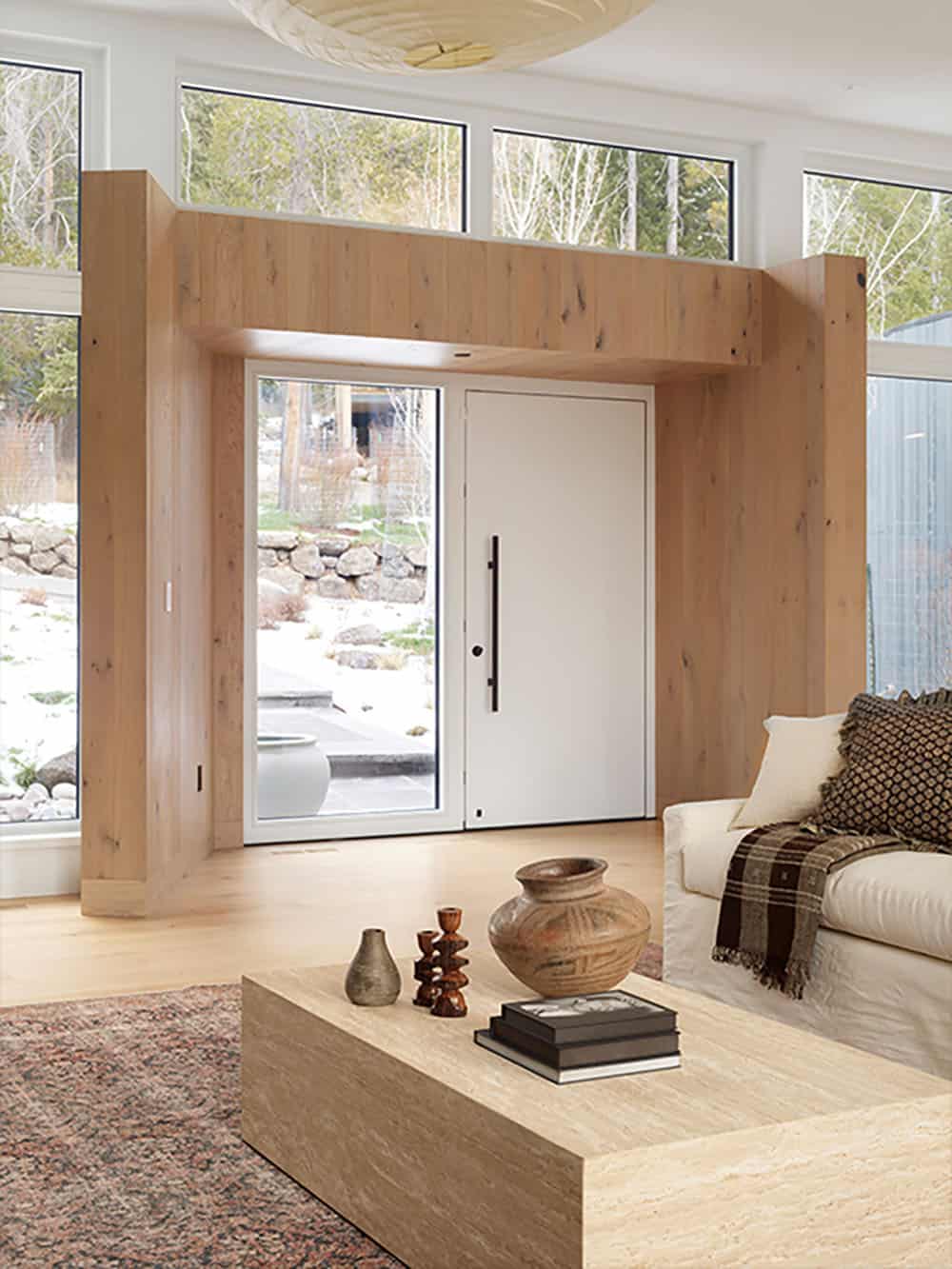 Entryway framed in white oak paneling with glass front door and travertine coffee table