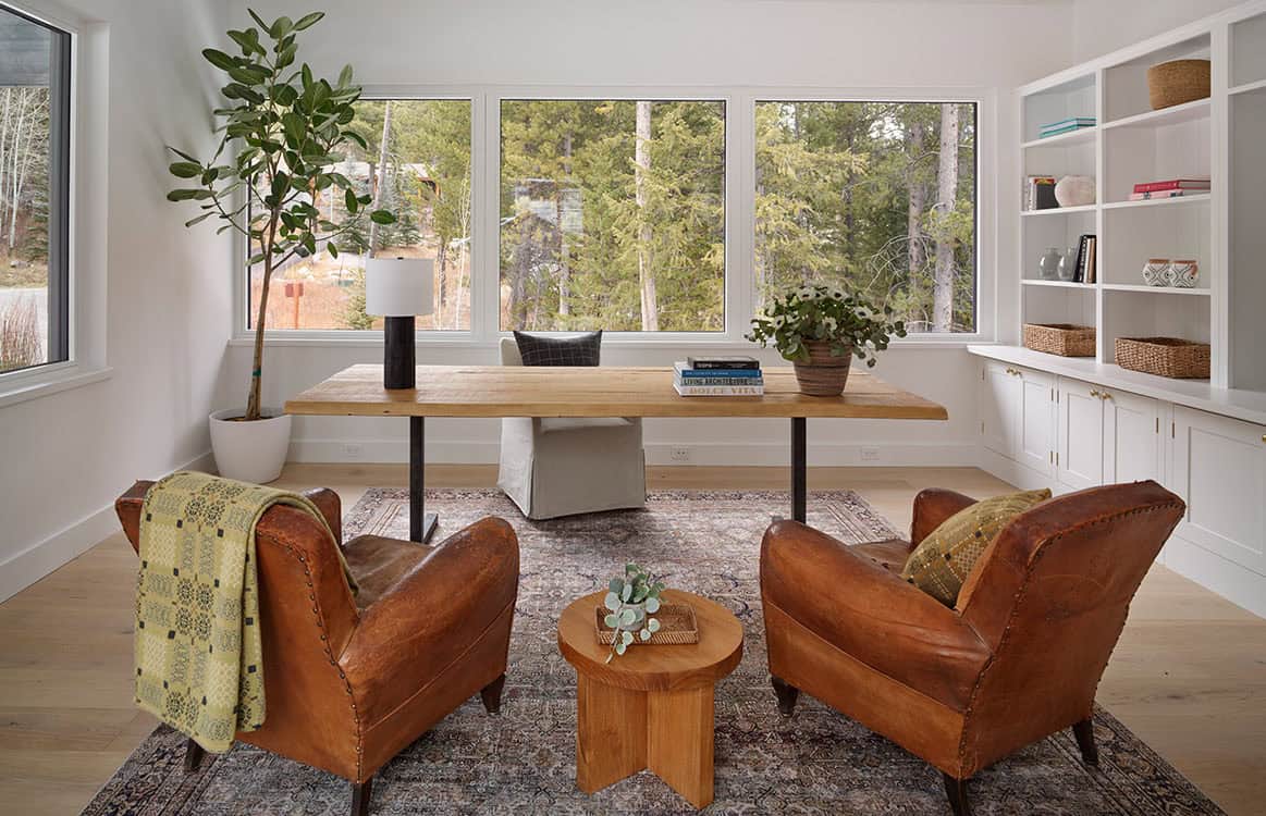 Home office with reclaimed wood desk, leather club chairs, and forest views through picture windows