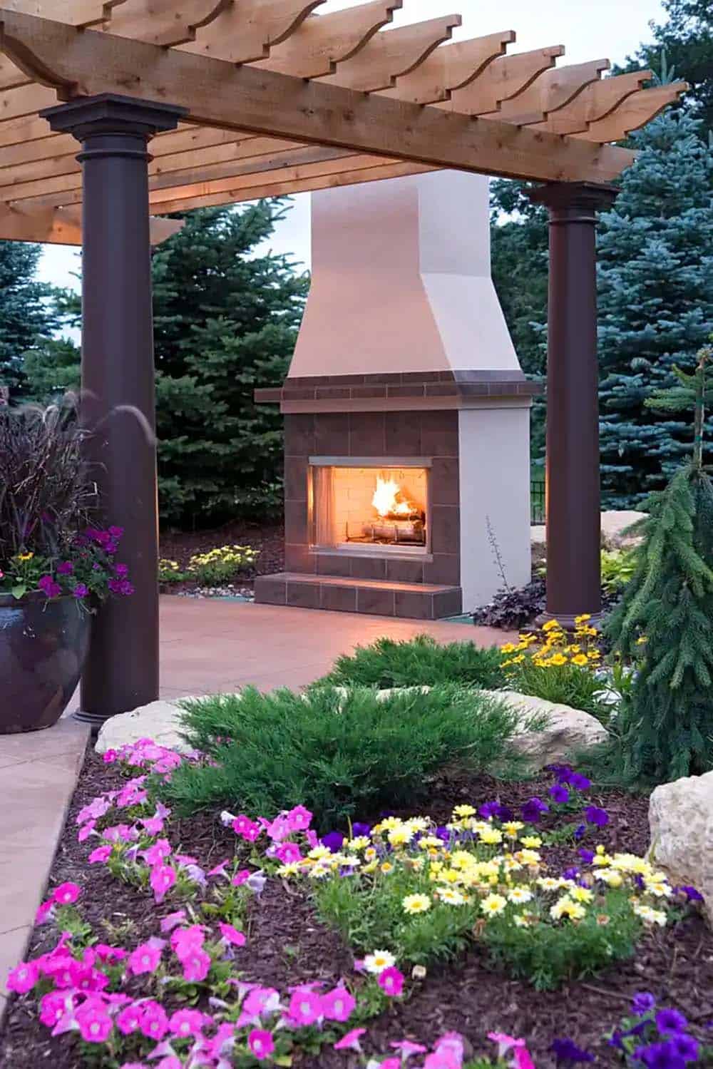 Lit outdoor fireplace under pergola surrounded by evergreen trees
