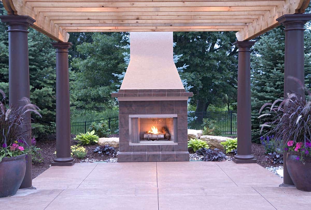 Outdoor stone fireplace lit at dusk beneath pergola columns