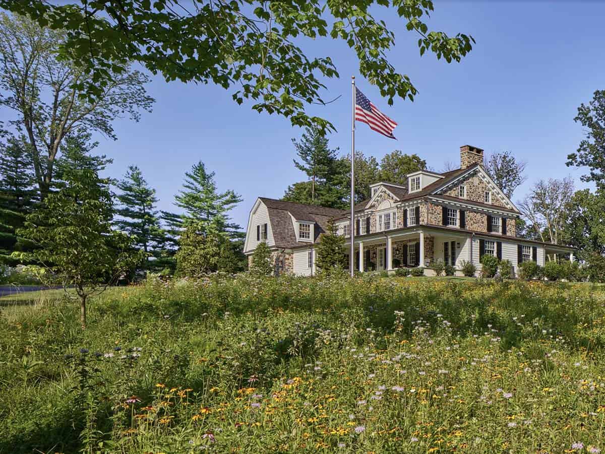 Stone-and-shingle farmhouse with gambrel roof seen across a wildflower meadow, American flag flying out front