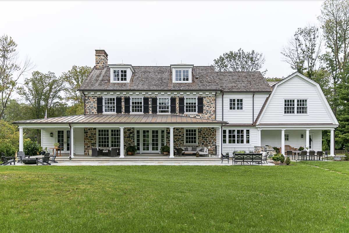 Rear facade with stone lower level, wraparound covered porch, standing-seam metal roof, and green lawn