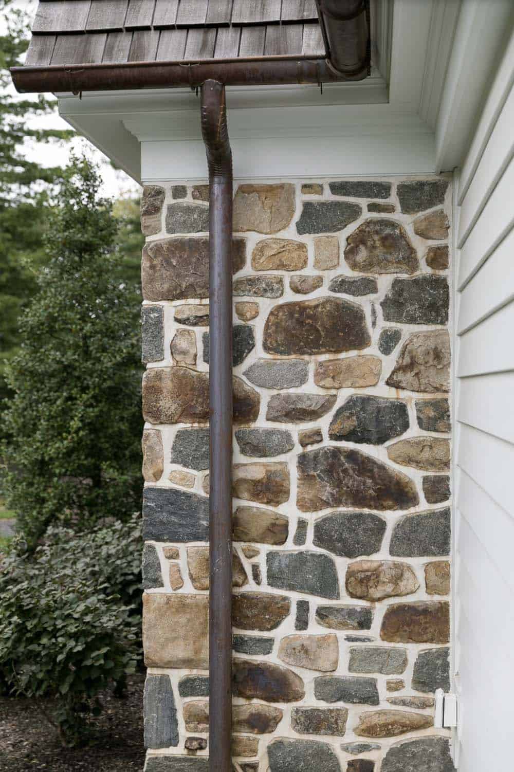 Fieldstone wall detail with copper downspout beneath a wood-shingled roof overhang