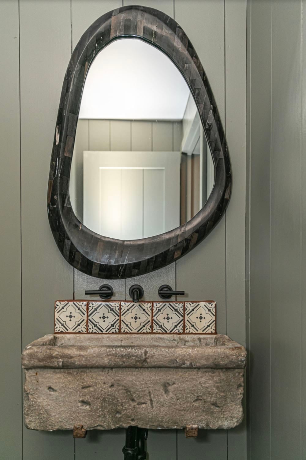Powder room vanity with organic-shaped inlaid mirror above a stone trough sink and patterned tile backsplash