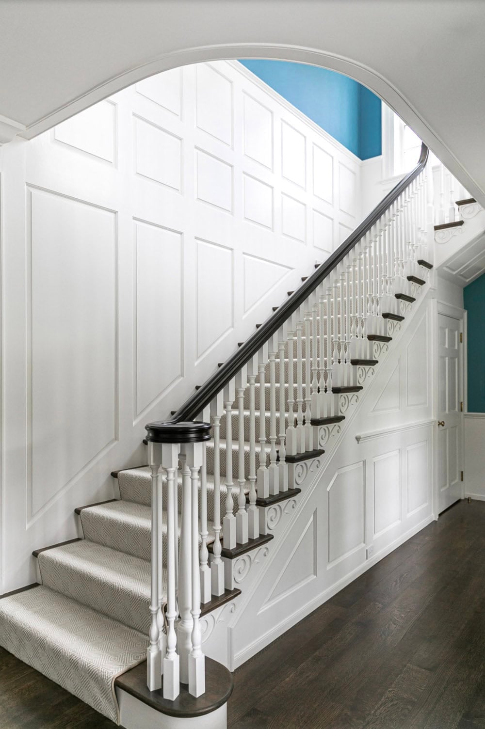 Grand staircase with white paneled wainscoting, dark wood handrail, and patterned runner