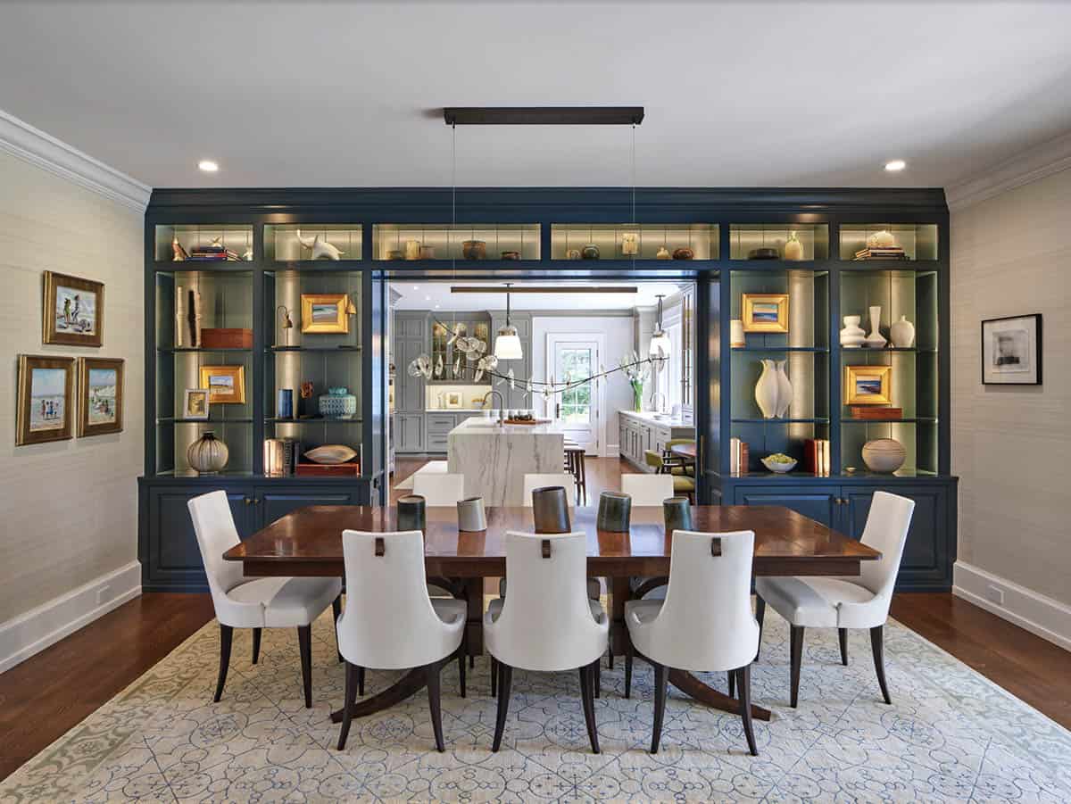 Dining room with navy built-in shelving, wood table, white chairs, opening to kitchen