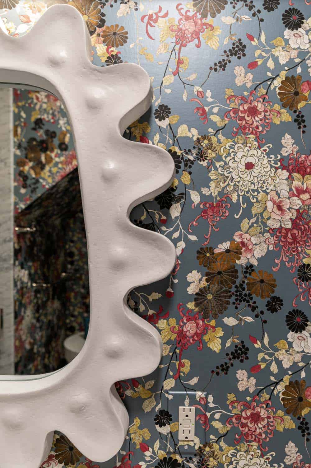 Sculptural white mirror frame against blue floral chinoiserie wallpaper in bathroom