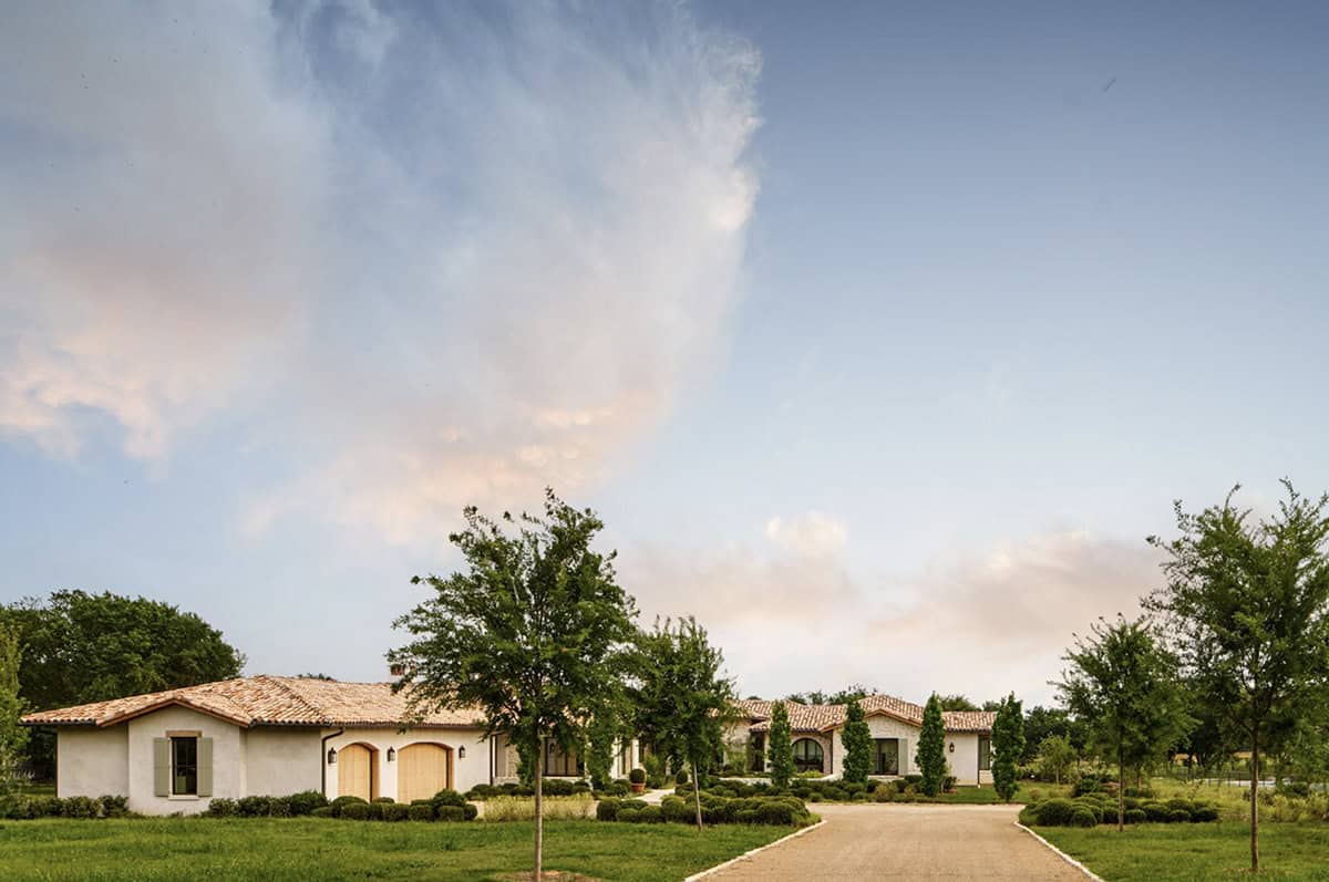 Mediterranean-style stone and stucco home with terracotta tile roof at dusk, viewed from the gravel driveway with young trees and clouds overhead