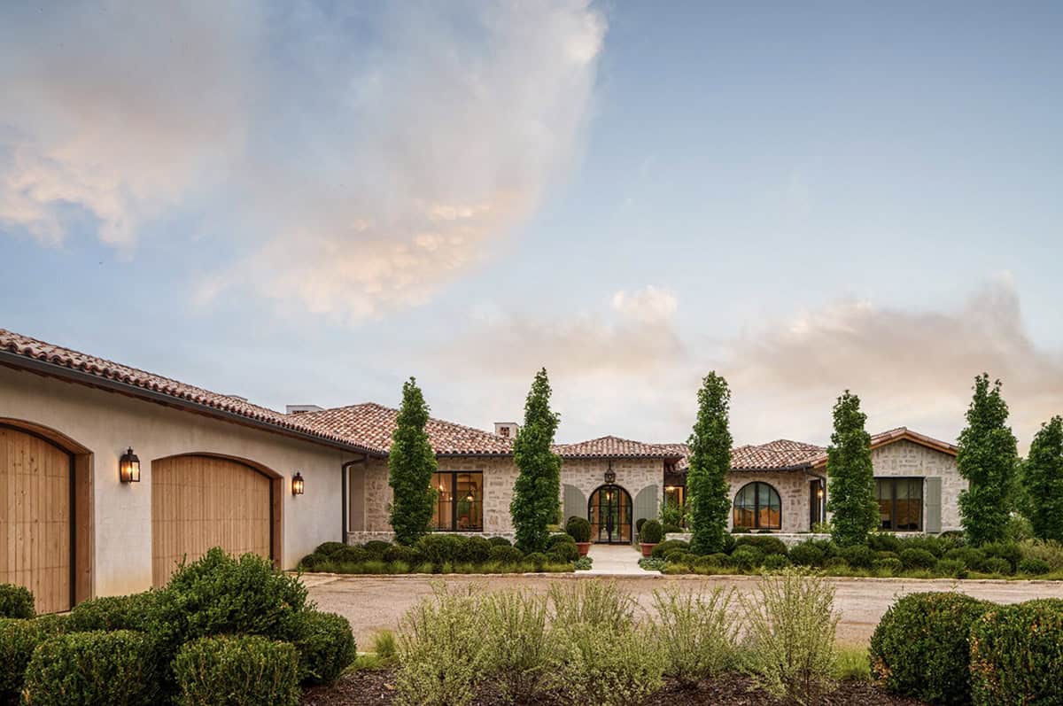 Front entrance of a Mediterranean stone house with arched wood garage doors, tall cypress trees, and boxwood landscaping
