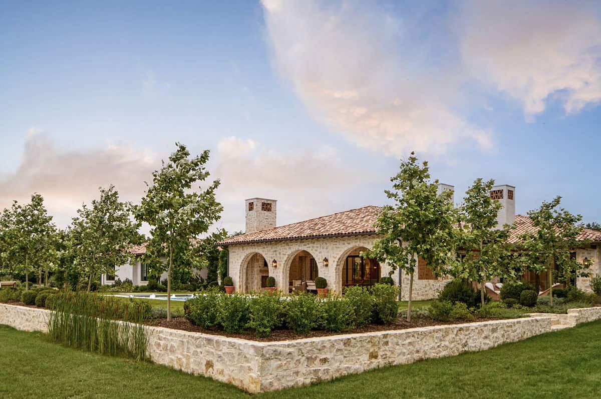Rear view of stone Mediterranean home with arched covered porch, chimneys, and pool glimpsed through manicured landscaping