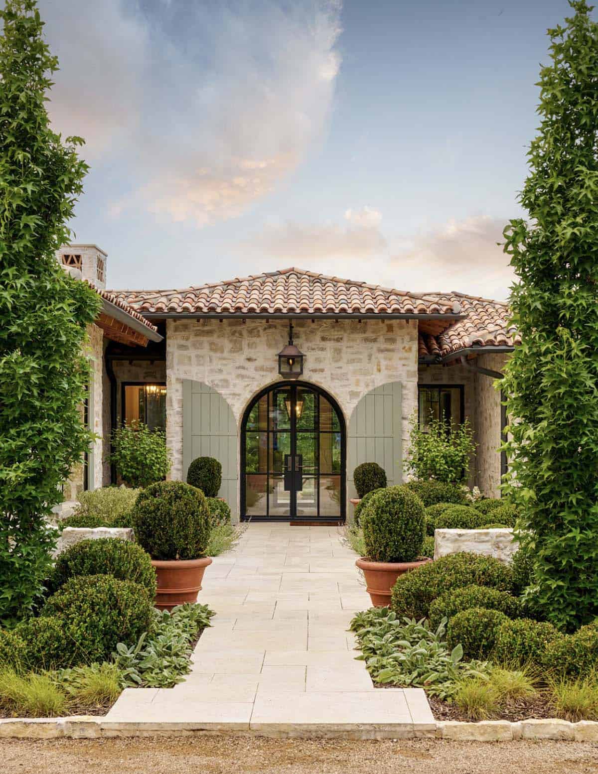 Arched black steel and glass front door flanked by climbing vines, boxwood topiaries in terracotta pots, and a stone pathway