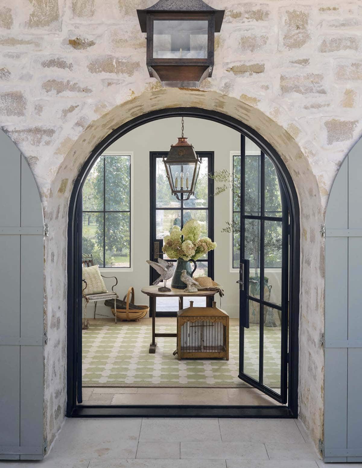 View through an open arched black steel door into a foyer with patterned green and white rug and rustic entry table