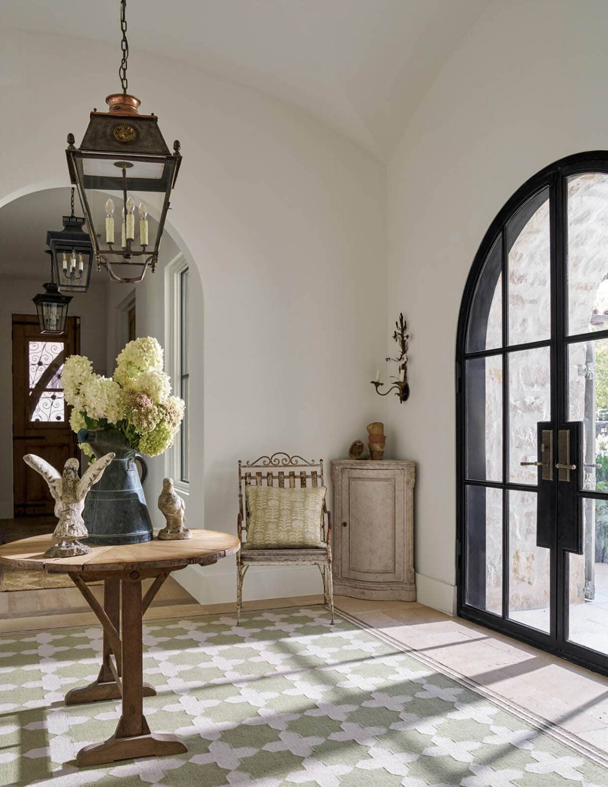 Bright foyer with arched black steel door, round wood table topped with pitcher of hydrangeas, iron chair, and patterned rug