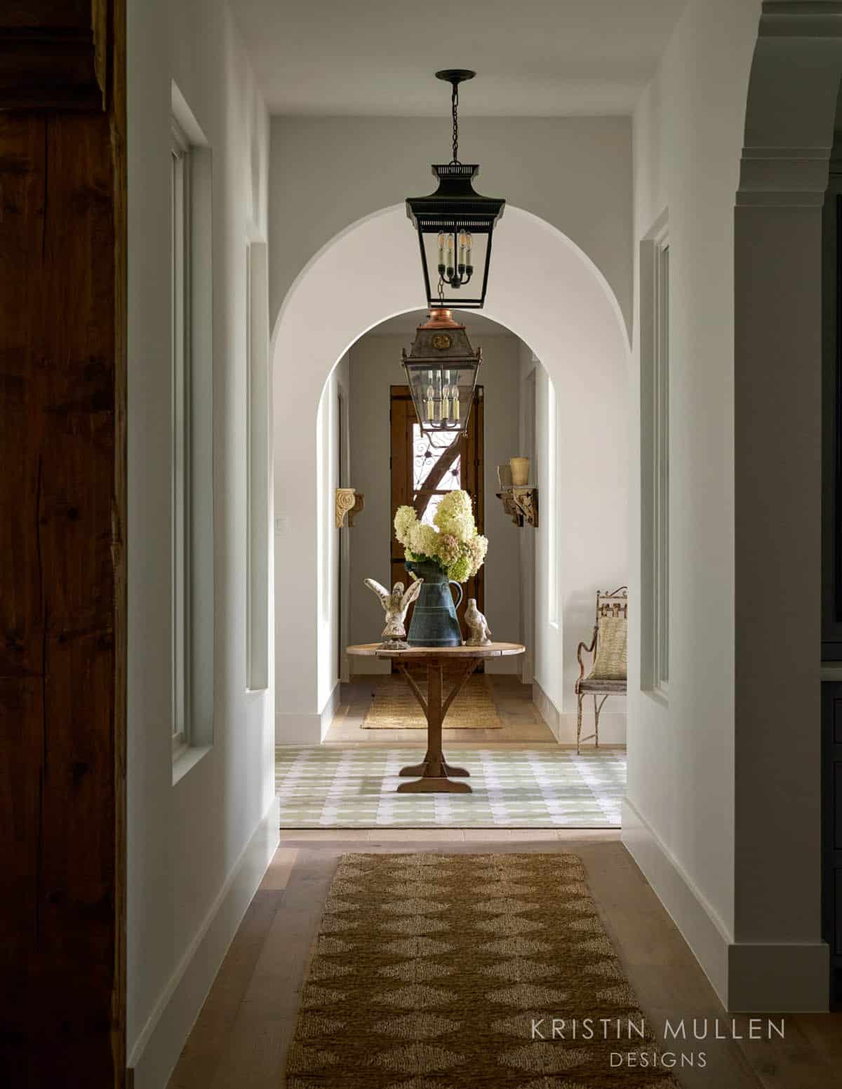 Hallway view through arched doorway toward a round entry table with hydrangeas, lanterns, and a jute runner