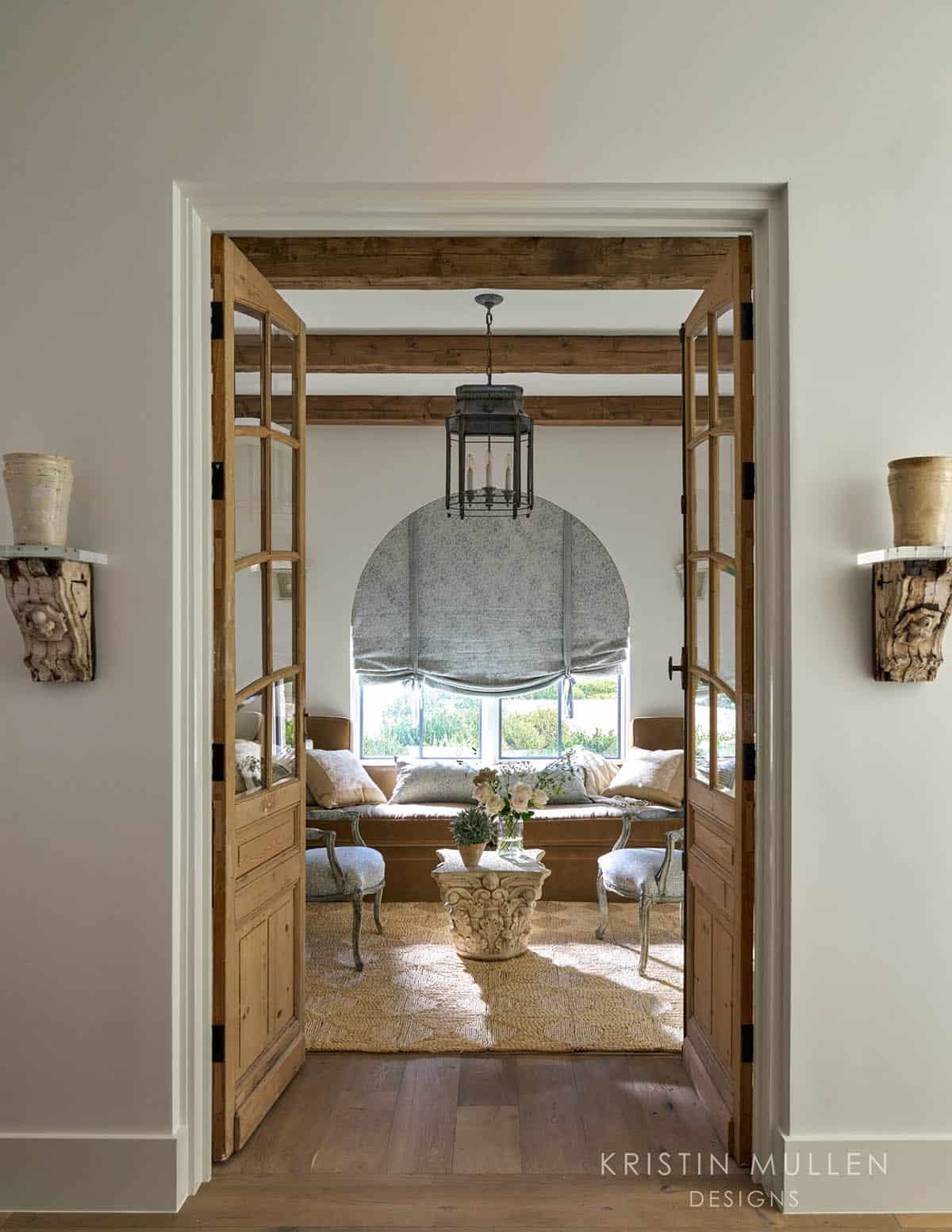 View through open pine French doors into a sunroom with a blue floral window seat and carved stone side table